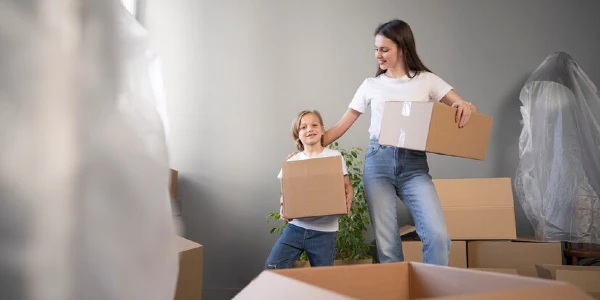Woman in white top and blue jeans holding a box standing with her kid holding a moving box.